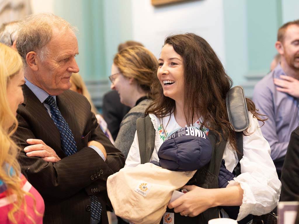 Female paediatrician with baby in a carrier speaking to colleagues at event
